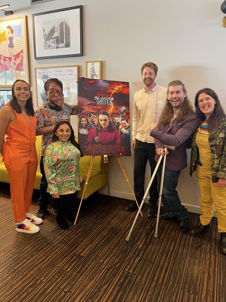 The leading cast members and director of Dragon Slayers United stand smiling round a large poster of the Film