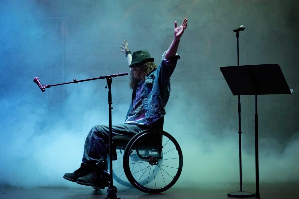 Dan sits in character in his wheelchair arms aloft wearing denim and a hat surround by smoke and mic and music stands in true rock star style