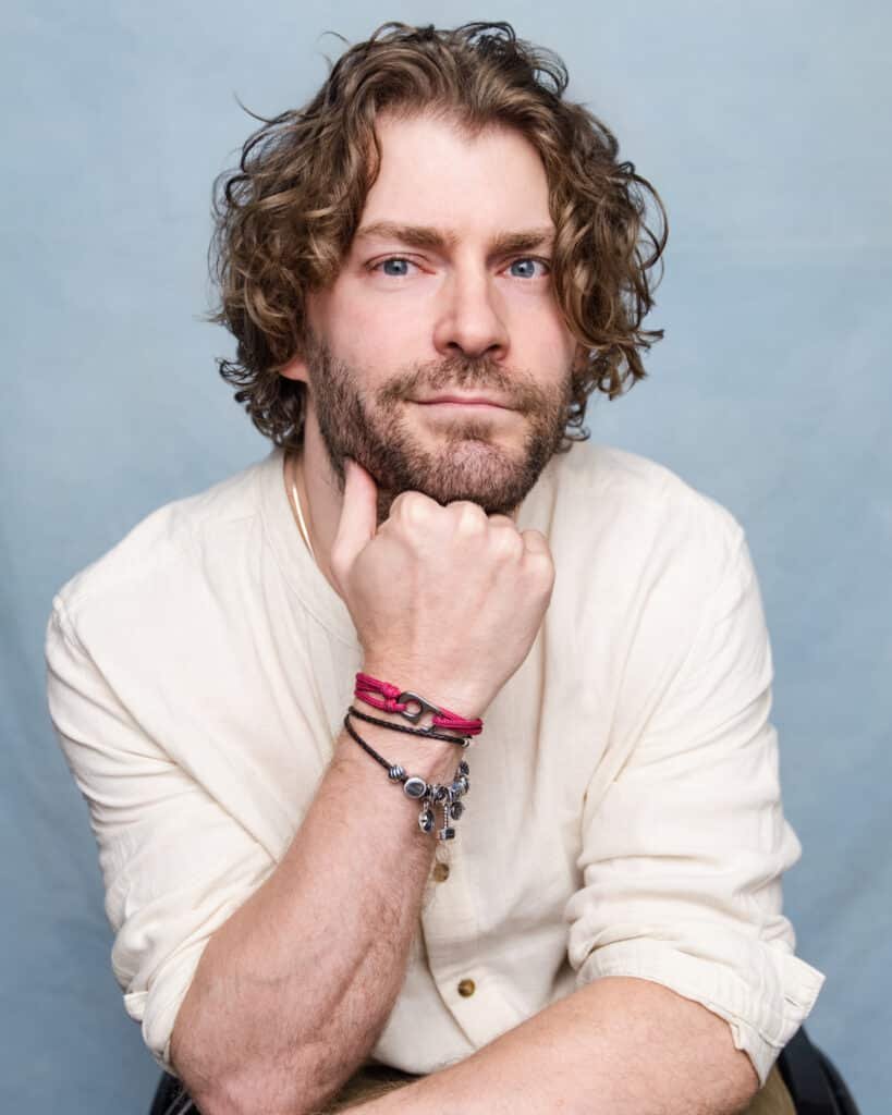 a picture of Dan Edge a white man with mid length curly dark blonde hair and a beard sits with his head rested on his hand in a pale shirt with a light blue background
