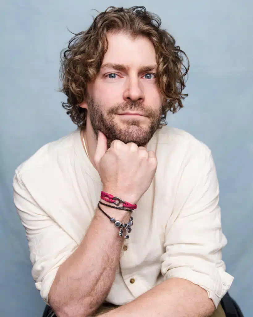 a picture of Dan Edge a white man with mid length curly dark blonde hair and a beard sits with his head rested on his hand in a pale shirt with a light blue background